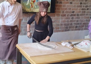 Zdjęcie ukazuje dziewczynkę oraz mężczyznę w czapkach kucharskich. Dziewczynka tnie ciasto na rogale przy użyciu szabli.