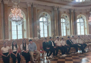 Uczniowie siedzą w Sali Tronowej na przepięknie zdobionych krzesłach. Z sufitu zwisają kryształowe żyrandole, a na podłodze znajduje się wpsaniały parkiet.