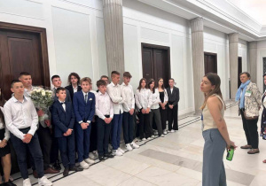 Uczniowie stoją na korytarzu łączącym Sejm z Senatem. Słuchają wykładu Pani przewodnik. Jeden z uczniów trzyma kwiaty, które zostaną wręczone w ramach podziękowania.