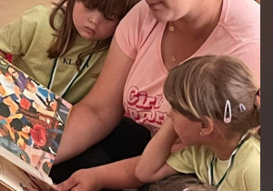 Dwie dziewczynki z grupy Motylki oglądają obrazki w książce którą w ręku trzyma mama jednej z nich i czyta.