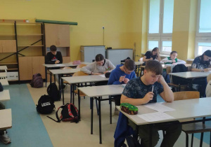 Zdjęcie ukazuje uczniów siedzących w klasowych ławkach, pochylonych nad kartkami z pytaniami konkursowymi.