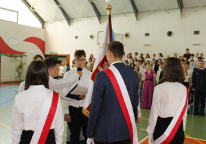 Uczniowie klasy VII przejmujący sztandar szkoły od uczniów klasy ósmej. W tle nauczyciele, goście zaproszeni, uczniowie.