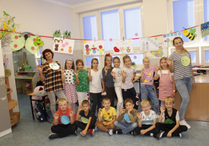 Grupa uczniów klasy drugiej wraz z nauczycielkami wykonują gest kółeczka dłońmi. Ruch ten nawiązuje do obchodów Międzynarodowego Dnia Kropki w naszej szkole. Dzieci mają na sobie ubrania w kolorowe kropki. Nad nimi są zawieszone prace plastyczne z kropek.