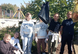 Na zdjęciu zadowoleni uczniowie z akcji. Dzieci trzymają worki z zebranymi odpadami. Wokół nich roślinność oraz plac zabaw.