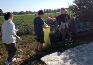 Na zdjęciu znajdują się uczniowie, którzy zbierają śmieci. Robią to na terenie wokół boiska szkolnego. Dzieci trzymają worki i wrzucają do nich zebrane śmieci.