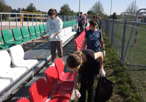 Grupa uczniów zbiera śmieci wokół trybun przy boisku szkolnym. Dzieci są zaangażowane w Akcję. Chętnie oraz dokładnie zbierają pozostawiane śmieci.