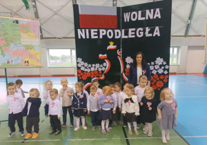 Grupa Pszczółki w strojach galowych z wychowawca na tle ścianki Wolna Niepodległa