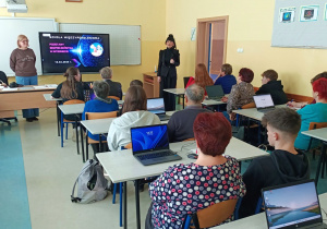 Sala lekcyjna. Pani Dyrektor wraz z nauczycielem informatyki wita uczestników spotkania „Szkoła Międzypokoleniowa”. Uczniowie i Seniorami siedzą przy stolikach, na których znajdują się włączone laptopy.