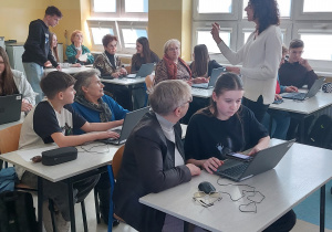 Sala lekcyjna, seniorzy wraz z wnukami siedzą w ławkach. Na ławkach znajdują się laptopy. Uważnie słuchają nauczycielki, która prowadzi wykład na temat bezpieczeństwa w Internecie.