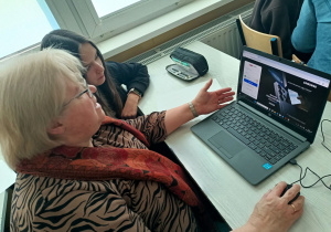 Babcia wraz ze swoją wnuczką siedzą przy stoliku, na którym znajduje się laptop. Pod czujnym okiem wnuczki, babcia zakłada konto e-mail.