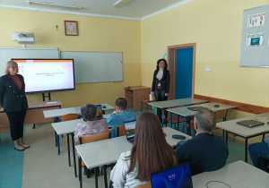 Sala lekcyjna. Pani Dyrektor i nauczyciel prowadzący zajęcia witają uczestników spotkania. Seniorzy wraz z uczniami siedzą przy stolikach na których leżą laptopy.