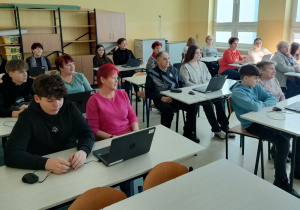 Sala lekcyjna, Seniorzy wraz z uczniami siedzą przy stolikach szkolnych na których leżą otwarte laptopy.