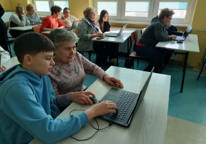 Seniorzy i uczniowie siedzą przy szkolnych stolikach, wybierając na laptopach różnorodne formy rozrywki online.