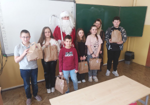 Uczniowie klasy czwartej, trzymając swoje prezenty, pozują do zdjęcia razem z nauczycielką języka polskiego i Świętym Mikołajem.