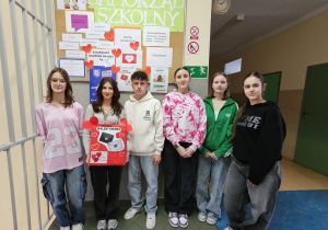 Grupa uczniów na tle tablicy samorządu szkolnego. Jedna z uczennic trzyma pudełko z pocztą walentynkową.