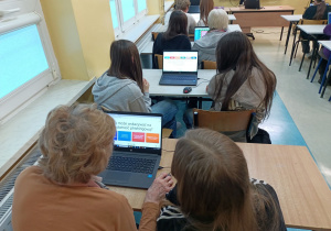 Sala lekcyjna, w której uczestniczki spotkania siedzą przy szkolnych stolikach i, korzystając z laptopów, wykonują ćwiczenia sprawdzające ich wiedzę na temat zagrożeń czyhających w internecie.