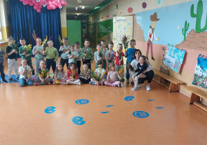 Grupa przedszkolna "Motylki" w szkolnym holu. W pierwszym rzędzie siedzą dziewczynki wraz z wychowawcą, a w drugim stoją chłopcy. Wszyscy trzymają własnoręcznie wykonane palemki wielkanocne.