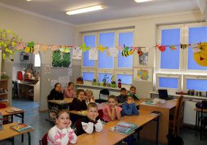 Sala lekcyjna z pracami dzieci rozwieszonymi na sznurku nad uczniami siedzącymi w ławkach.