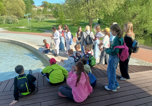 Park Centralny w Olsztynie. Uczestnicy wycieczki odpoczywają przy fontannie – część uczniów siedzi na drewnianym podeście i murku przy wodzie, pozostali stoją w pobliżu. Wokół teren zielony na którym rosną krzewy i drzewa.