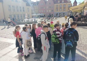 Uczniowie zwiedzają Stare Miasto w Olsztynie. Po lewej stronie znajduje się budynek biblioteki, przed nim fontanna z wypływającą wodą, w oddali zabytkowe kamienice.