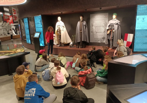 Uczniowie siedzą na podłodze w Muzeum w Grunwaldzie. Słuchają pani Przewodnik, która omawia ubiory i wyglądy braci zakonnych.