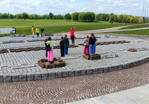 Makieta wykonana z kolorowych kamieni, przedstawiająca rozstawienie wojsk i ich obozów przed bitwą pod Grunwaldem. Uczniowie stoją na makiecie i słuchają pani Przewodnik, która opowiada o strategicznym układzie sił przed historycznym starciem.