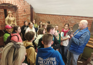 Uczniowie zwiedzają wystawę etnograficzno-historyczną. Z zaciekawieniem słuchają przewodnika, który prezentuje eksponaty z epoki średniowiecza, przybliżając codzienne życie i kulturę dawnych czasów.