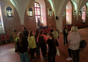 Uczniowie zwiedzają salę rycerską w Muzeum Ziemi Nidzickiej. Przewodnik opowiada o historii regionu i prezentuje kopie uzbrojenia piechoty chłopskiej oraz rycerstwa z okresu bitwy pod Grunwaldem.