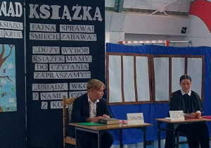 Sędzia i oskarżyciel siedzą przy stolikach. Ochroniarz stoi przy drzwiach. Na tablicy dekoracyjnej znajduje się plakat pt. ,,Tysiąc powodów, by czytać” oraz hasła zachęcające do czytania książek. Dekoracją jest również parawan będący tłem sali sądowej.