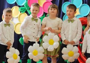 Trzech chłopców i dziewczynka z balonowymi stokrotkami pozują do zdjęcia na tle dekoracji z okazji Dnia Mamy i Taty.