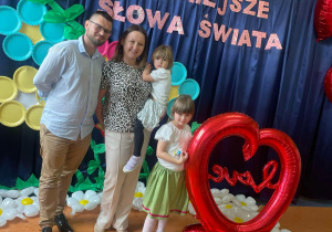 Rodzice wraz dziewczynkami z grupy Motylki i Biedronki na tle dekoracji z okazji dnia Mamy i Taty przy dużym sercowym balonie.