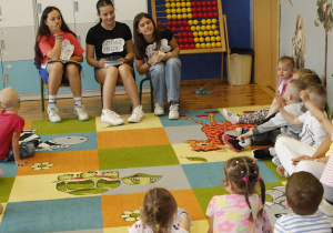 Uczennice z klasy VII b i z klasy VI b rozmawiają z przedszkolakami z grupy ,,Biedronki”. Dzieci siedzą na dywanie. Razem z nimi siedzi pomoc nauczyciela wychowawcy.