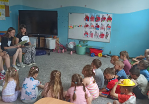 Uczennica z klasy VI a czyta przedszkolakom z grupy ,,Motylki” wiersz Juliana Tuwima ,,Słoń Trąbalski”. Obok niej na krzesełkach siedzą uczennice z klasy VI a i z klasy VI b. Przedszkolaki siedzą na dywanie z wychowawcą grupy.