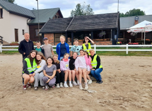 Grupowe zdjęcie z wycieczki – na pierwszym planie siedzą dziewczynki razem z wychowawczynią, za nimi stoją chłopcy wraz z nauczycielem. Wszyscy uśmiechnięci, pozują do wspólnej fotografii.