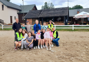 Grupowe zdjęcie z wycieczki – na pierwszym planie siedzą dziewczynki razem z wychowawczynią, za nimi stoją chłopcy wraz z nauczycielem. Wszyscy uśmiechnięci, pozują do wspólnej fotografii.