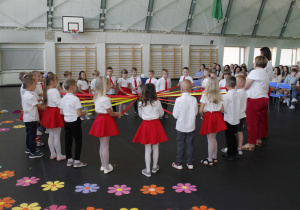 Dzieci kończące odział przedszkolny z grupy Krasnale i Misie z wychowawcami w kole trzymając na zmianę w dłoniach wstążkę w kolorze czerwonym lub żółtym