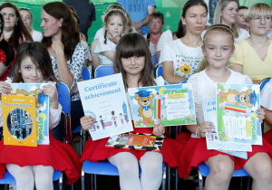 Dziewczynki z grupy Misie siedzą na krzesełkach w dłoniach trzymają dyplom ukończenia oddziału przedszkolnego, dyplom ukończenia zajęć z języka angielskiego oraz książki w tle rodzice biorący udział w uroczystości zakończenia przedszkola.