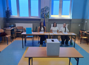 Trzy dziewczyny w komisji wyborczej siedzą przy stoliku, na środku sali znajduje się na stoliku urna do wrzucania głosów.