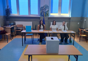 Trzy dziewczyny w komisji wyborczej siedzą przy stoliku, na środku sali znajduje się na stoliku urna do wrzucania głosów.