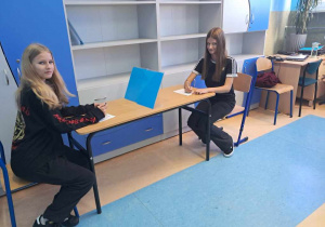 Dwie Uczennice z klasy 8 a siedzą przy stole na przeciwko siebie i wypełniają kartę do głosowania na środku stolika znajduje się pleksa.