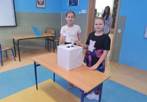 Dwie uczennice z klasy 4 stoją przy urnie i wrzucają głosy do skrzynki.