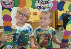 Dwóch chłopców z grupy Słoneczka pozują do zdjęcia z ozdobną ramką z okazji Dnia Kropki w własnoręcznie zrobionych koszulkach w kropki.