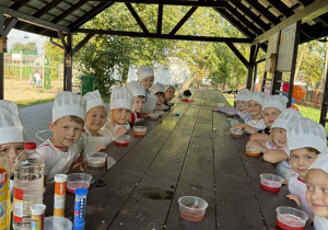 Dzieci z Grupy Żabki ubrane w białe zestawy kucharskie podczas doświadczeń w drewnianej altanie.