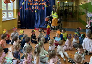 Pani Jesień wjeżdża do oddziału przedszkolnego na rowerze w kasku na głowie i kamizelce odblaskowej machając do dzieci.