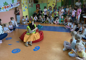 Pani Jesień siedzi w środku koła na czerwonej poduszce w ręku trzyma kask rowerowy, opowiada dzieciom o zasadach bezpieczeństwa w czasie jazdy rowerem.