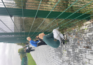 Uczniowie zbierają śmieci przy ogrodzeniu boiska szkolnego.