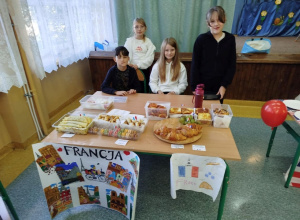 Grupa uczniów klasy 4 prowadzi stoisko kulinarne poświęcone Francji. Na stole znajdują się croissanty, makaroniki, naleśniki, zapiekanki i inne francuskie wypieki. Przed stołem wisi plakat z
