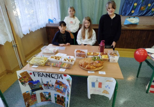 Grupa uczniów klasy 4 prowadzi stoisko kulinarne poświęcone Francji. Na stole znajdują się croissanty, makaroniki, naleśniki, zapiekanki i inne francuskie wypieki. Przed stołem wisi plakat z napisem ‘Francja’, z kolorowymi rysunkami Paryża oraz francuską flagą.