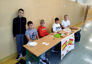 Grupa uczniów klasy 4 prowadzi stoisko kulinarne poświęcone Hiszpanii. Na stole znajdują się hiszpańskie przysmaki, takie jak ślimaczki. Przed stołem wisi plakat z napisem "HISZPANIA" oraz flagą Hiszpanii.