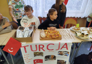 Klasa 6 prowadzi stoisko kulinarne poświęcone Turcji. Na stole leżą kebaby owinięte w folię, pide, wrapy i kanapki. Przed stołem wisi plakat z napisem ‘Turcja’ oraz zdjęciami tureckich potraw i flagą Turcji.